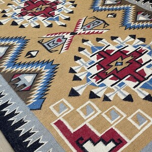 May include: A tan-colored woven rug with geometric patterns in red, white, blue, and black. The rug features repeating diamond and zigzag designs, with a fringed edge. The overall style is reminiscent of Southwestern or Native American textiles.