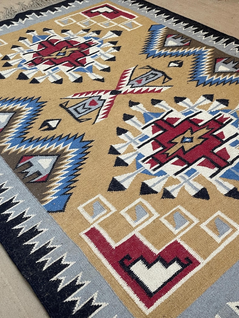 May include: A tan-colored woven rug with geometric patterns in red, white, blue, and black. The rug features repeating diamond and zigzag designs, with a fringed edge. The overall style is reminiscent of Southwestern or Native American textiles.