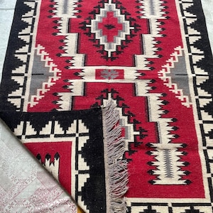 May include: A red, black, and white wool rug with a geometric pattern. The rug has fringe on all four sides.