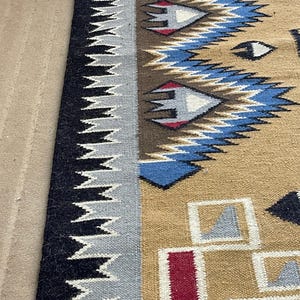 May include: A woven rug with a geometric pattern. The rug features a tan background with blue, red, and white geometric shapes. A black and gray zig-zag border runs along the edge of the rug. The rug is likely made of wool or a similar material.