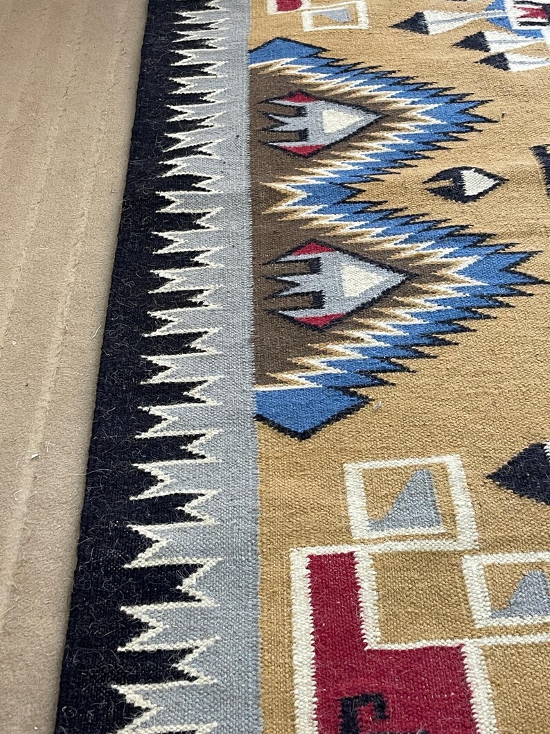 May include: A woven rug with a geometric pattern. The rug features a tan background with blue, red, and white geometric shapes. A black and gray zig-zag border runs along the edge of the rug. The rug is likely made of wool or a similar material.