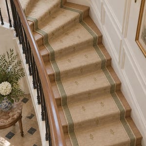 May include: A staircase runner featuring a light beige textured fabric with a repeating small leaf pattern and sage green borders. The runner is installed over wooden steps, complemented by a dark wood handrail and black metal balusters.