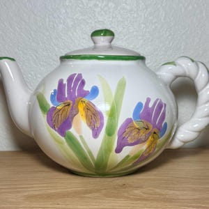 May include: White ceramic teapot with a lid and a twisted handle. The teapot is decorated with purple, yellow, and blue iris flowers and green leaves. The lid has a green rim and a pink knob.