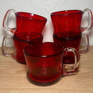 Puede incluir: Cinco tazas de vidrio rojo con asas transparentes apiladas sobre una superficie de madera. Las tazas tienen un diseño curvo. Las tazas son de color rojo intenso y parecen ser vintage o retro.