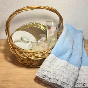 Puede incluir: Una cesta dorada tejida contiene una pequeña taza y platillo blancos, un espejo con borde dorado y dos vasos con diseños florales rosas. Una toalla azul claro con ribete de encaje blanco está drapeada a un lado. La escena está sobre una superficie de madera clara.