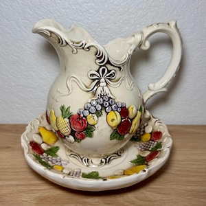 Könnte beinhalten: Cremefarbener Keramikkrug und passendes Untertassen-Set. Der Krug hat einen dekorativen Griff und Ausguss, mit einem detaillierten Relief einer Obstgirlande. Die Untertasse spiegelt das Fruchtdesign wider. Das Set ist wahrscheinlich Vintage.