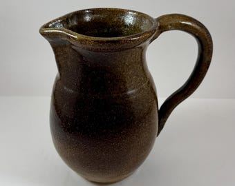 May include: A handcrafted, brown ceramic pitcher with a rounded body and a curved handle. The pitcher has a spout for easy pouring and a speckled texture, suggesting a rustic or artisanal aesthetic. The color is a deep, earthy brown.
