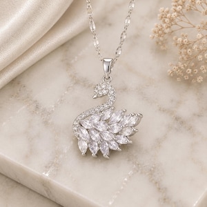 Può includere: Una collana con pendente a forma di cigno argentato. Il cigno è tempestato di cristalli trasparenti e ha una catena delicata. Il corpo del cigno è fatto di cristalli a forma di marquise. La collana è esposta su una superficie di marmo.