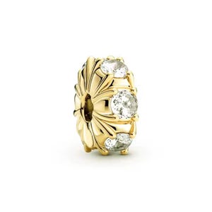 May include: A gold-colored charm with three clear, round gemstones. The charm has a floral design with a central hole. The gemstones are evenly spaced around the charm, adding sparkle and detail to the jewelry.