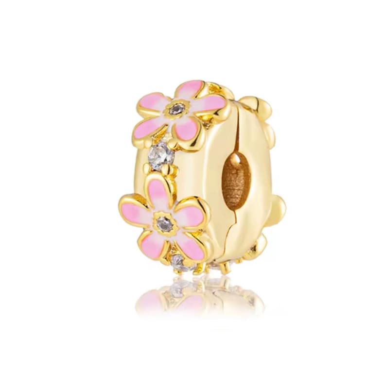 May include: A gold-toned charm bead featuring a central band decorated with pink enamel flower petals and small clear stones. The cylindrical bead has a polished metallic finish and a hollow center, designed to slide onto a bracelet or necklace chain.