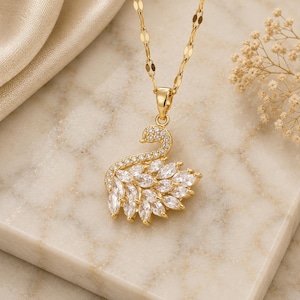 Può includere: Una collana con pendente a forma di cigno dorato. Il cigno è ornato di cristalli trasparenti e scintillanti. La catena è delicata. La collana è esposta su una superficie di marmo.