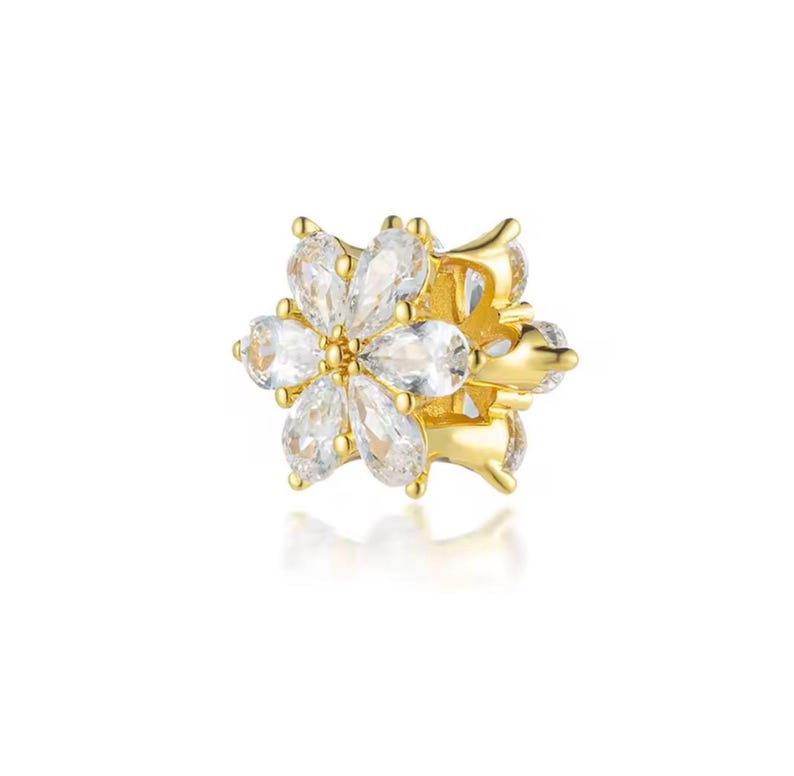 May include: A gold-colored floral-shaped charm with clear, teardrop-shaped gemstones. The charm has a detailed design with multiple petals and a central setting, creating a sparkling, elegant appearance. The charm is likely for jewelry.