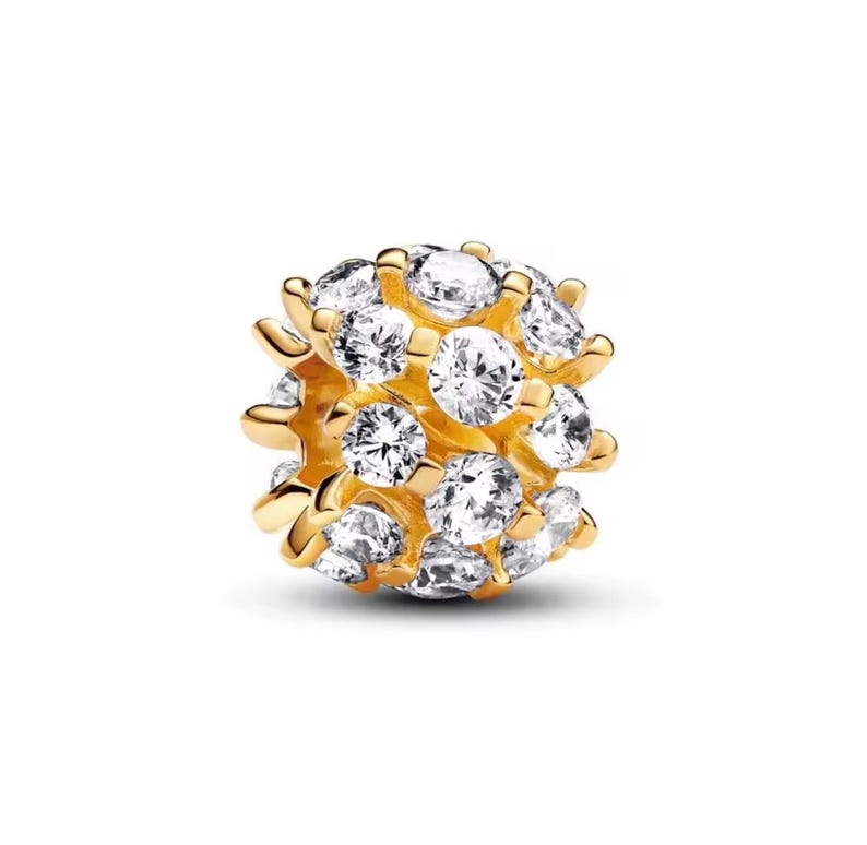 May include: A gold-colored charm with a cluster of clear, round-cut gemstones. The charm has a spherical shape, with the gemstones closely set together. The gold setting has a polished finish, and the gemstones sparkle brightly against the gold.