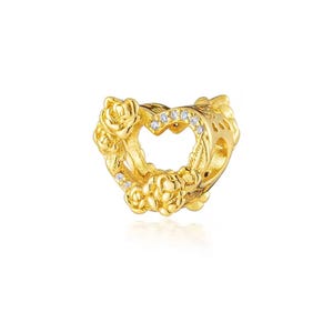 May include: A gold-colored charm with a heart-shaped cutout. The charm is adorned with rose and leaf details, and small, clear gemstones are set around the heart. The charm is designed to be a pendant or bead.