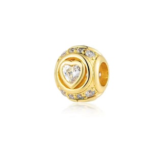 May include: A gold-colored charm with a heart-shaped gemstone at the center. The charm is round with smaller gemstones encircling the heart and around the sides. The charm has a hole on the side for stringing.