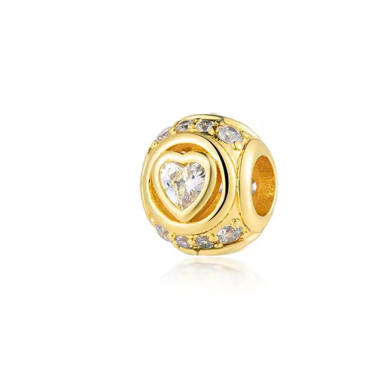 May include: A gold-colored charm with a heart-shaped gemstone at the center. The charm is round with smaller gemstones encircling the heart and around the sides. The charm has a hole on the side for stringing.