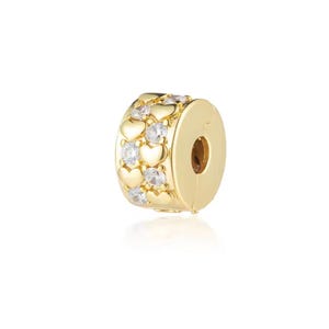 May include: A gold-colored charm with a cylindrical shape. The charm is decorated with alternating rows of small hearts and clear, round gemstones. The charm has a hole on one side, likely for threading onto a bracelet or necklace.