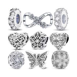 May include: A selection of silver charm beads in various designs. The charms include an infinity symbol with floral details, heart shapes, a butterfly, and beads with clear crystal embellishments. Some charms have the word "LOVE" on them.