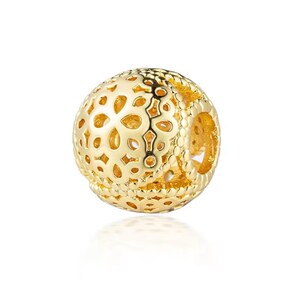 May include: A spherical gold charm bead featuring an intricate openwork filigree pattern with floral-inspired cutouts and a beaded border design. The surface has a polished metallic finish with a hollow center for threading onto a bracelet.