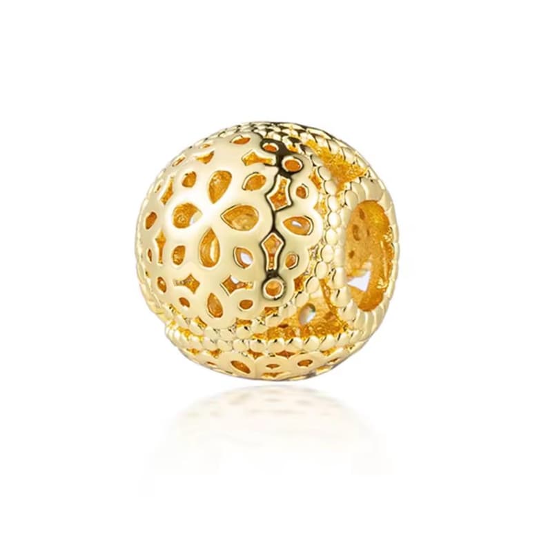 May include: A spherical gold charm bead featuring an intricate openwork filigree pattern with floral-inspired cutouts and a beaded border design. The surface has a polished metallic finish with a hollow center for threading onto a bracelet.