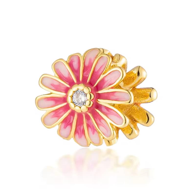 May include: A gold-toned metal charm shaped like a flower with layered petals featuring a gradient of pink and white enamel. The center of the flower is decorated with a small, clear, round faceted stone surrounded by a textured gold-toned border.