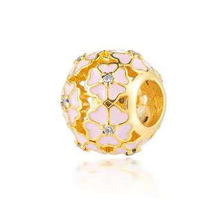 May include: A spherical gold charm bead featuring an openwork design of interconnected flowers with light pink enamel petals and a small clear crystal set in the center of each flower. The bead has a large circular opening through the middle.