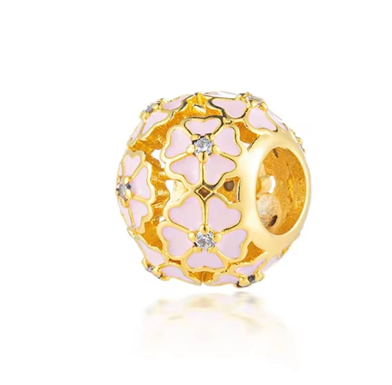 May include: A spherical gold charm bead featuring an openwork design of interconnected flowers with light pink enamel petals and a small clear crystal set in the center of each flower. The bead has a large circular opening through the middle.
