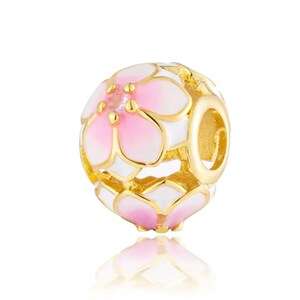 May include: A spherical charm bead featuring a gold-tone metal frame with white and pink enamel flower petal designs. The bead has a central opening for threading onto a bracelet or necklace chain, showcasing a detailed floral pattern throughout.