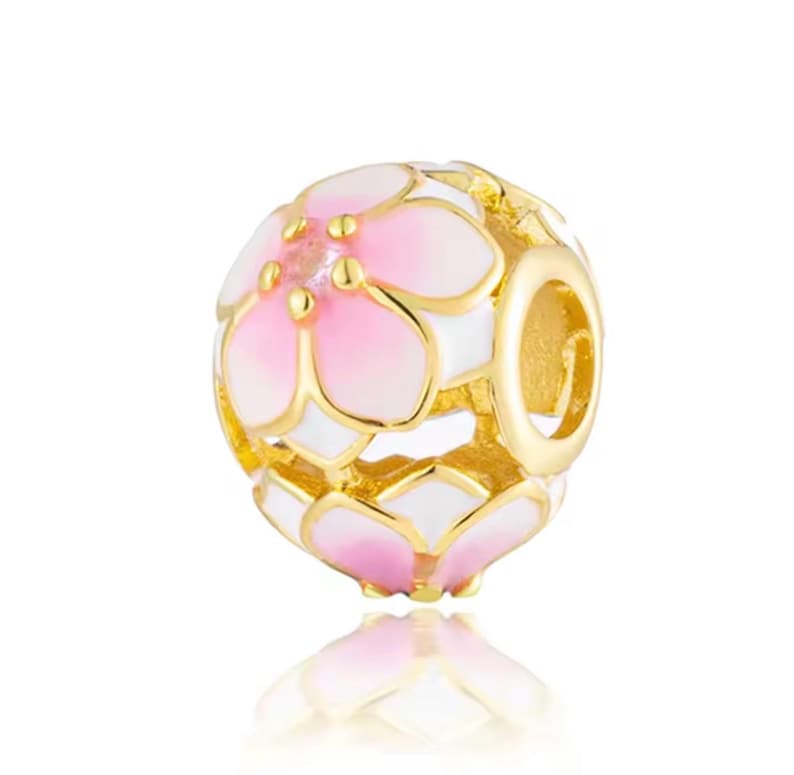 May include: A spherical charm bead featuring a gold-tone metal frame with white and pink enamel flower petal designs. The bead has a central opening for threading onto a bracelet or necklace chain, showcasing a detailed floral pattern throughout.