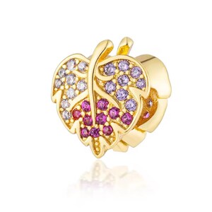 May include: A gold-toned charm shaped like a leaf, featuring a central vein and embedded with small, round, multi-colored gemstones. The stones are arranged in distinct sections, displaying clear, light purple, and vibrant pink hues across the surface.