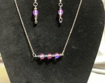 May include: A silver chain necklace with a bar pendant featuring four iridescent purple glass beads. Matching earrings with a single iridescent purple glass bead dangle from silver hooks.