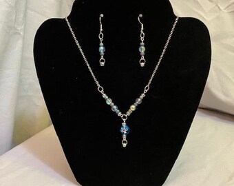 May include: A silver necklace with iridescent blue and green beads and a blue bead with a white flower design. Matching earrings with iridescent blue and green beads.