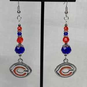 May include: A pair of dangle earrings featuring a silver-toned football-shaped charm with an orange and white design. The earrings have red and blue beads and silver-toned hooks.