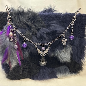 May include: A dark blue and black faux fur handbag adorned with a silver-toned chain featuring purple glitter beads, a silver skull charm, a bat-wing pendant, and a teardrop-shaped crystal with a white cross symbol, accented by purple feathers.
