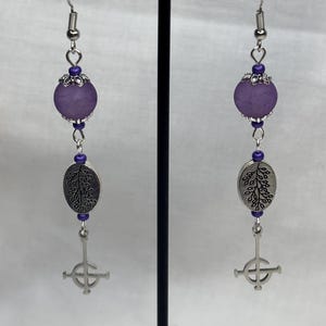 May include: A pair of dangle earrings with a purple bead, silver-colored leaf charm, and a cross pendant. The earrings are displayed on a black stand against a white background. The earrings have a hook closure.