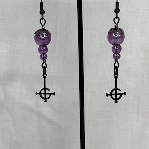 May include: A pair of black metal earrings with a gothic cross pendant. The earrings feature a series of purple beads and a large, round purple bead at the top. The earrings are displayed on a black stand.