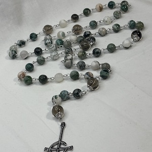 May include: A rosary with alternating green, white, and brown speckled beads strung on silver-colored chain. The rosary features a silver-colored pendant with a stylized cross design at the bottom.