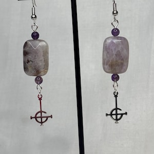 May include: A pair of earrings featuring rectangular, light purple gemstones with silver-tone metal hooks. Each earring has a small purple bead and a dangling, stylized cross pendant. The earrings are displayed on a black stand.