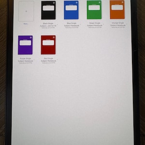 Peut inclure: Un écran de tablette affiche des modèles de cahiers. Les modèles comprennent des cahiers noirs, bleus, verts, oranges, violets et rouges. Chaque cahier a un rectangle blanc et une étoile. L'écran affiche également l'heure et la date.