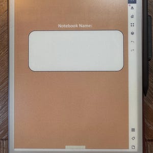 May include: A digital notebook with a brown screen and a white rectangular area for a notebook name. A black stylus is attached to the side. The notebook is on a wooden surface.