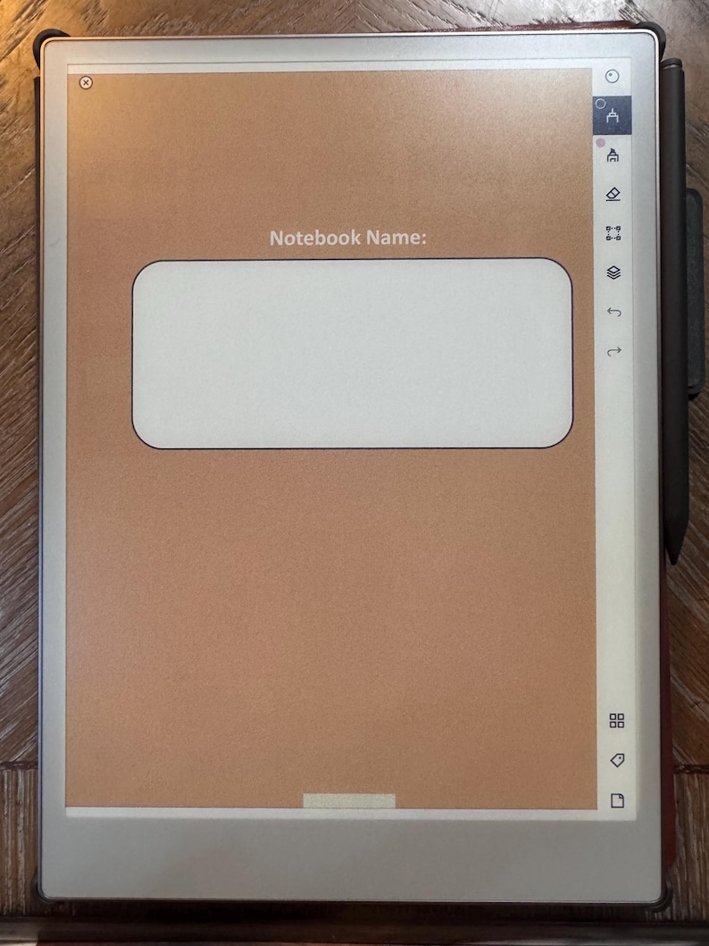 May include: A digital notebook with a brown screen and a white rectangular area for a notebook name. A black stylus is attached to the side. The notebook is on a wooden surface.
