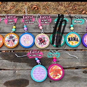 May include: A collection of nine circular car air fresheners featuring colorful graphic designs and text. Each ornament includes a hanging string and two decorative beads. Designs include phrases like Anti Social Cowgirls Club, Space Cowgirl, Mama, and Love Yourself More.
