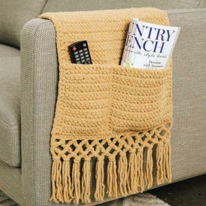 May include: A mustard yellow crochet sofa armrest tidy with two pockets and a fringe trim. A remote control and a magazine titled "Country French" are in the pockets. The tidy is draped over the arm of a grey sofa.