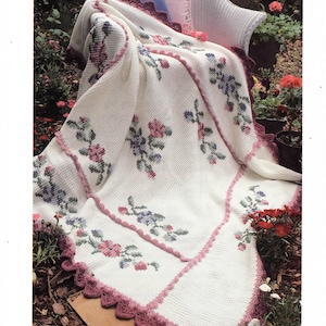 Puede incluir: Una manta blanca de ganchillo con bordados florales en rosa, morado y verde. La manta tiene un borde festoneado en un color rosa más oscuro y está sobre una silla de mimbre blanca. Se ven cojines decorativos.