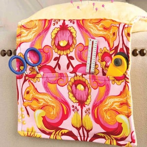May include: A pink, yellow, and red fabric sewing organizer with a floral and squirrel pattern. The organizer has pockets holding blue scissors, a ruler, and a yellow rotary cutter. Pins are stuck in the yellow top.