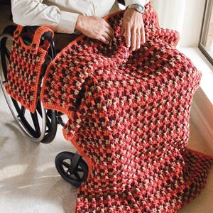 May include: A handmade crochet blanket in shades of red, orange, and beige. The blanket has a textured pattern and is draped over a person's lap. A matching crochet bag is attached to a wheelchair.
