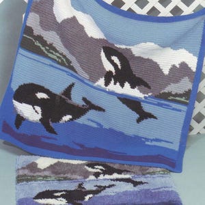 Pode incluir: Um cobertor de crochê azul e branco com uma cena de orcas. O cobertor mostra orcas nadando no oceano com montanhas ao fundo. O cobertor tem uma borda azul e um tapete menor correspondente.