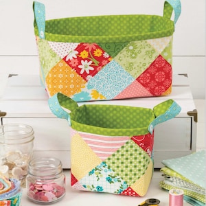 May include: Two handmade fabric storage baskets. The baskets are rectangular with green polka dot lining and handles. The exterior is patchwork with floral and geometric patterns in various colors, including red, yellow, and blue. The baskets are on a white surface.