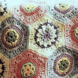 May include: Close-up of a handmade crochet blanket. The blanket features a repeating pattern of hexagonal shapes in various colors, including cream, pink, gray, and purple. The center of each hexagon has a floral design. The text "Beessia Vintage Collection" is visible.