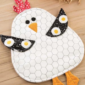 Pode incluir: Porta-panela branco em forma de frango com desenhos de ovos pretos e brancos nas asas. O porta-panela tem um padrão de arame de frango preto e branco e pés laranja.